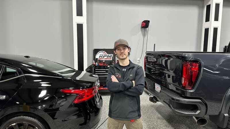 Jon Bobillier, paintless dent repair technician at RPM Dent Repair in Raynham, Massachusetts, trained by master technician John Scatto Jr., standing professionally in the PDR shop between customer vehicles with the RPM Dent Repair logo visible in the background
